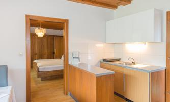 A modern kitchen corner with wooden surfaces and a dining table. In the background, a bedroom with a double bed can be seen.