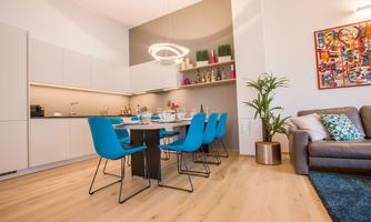 A modern living room with a large dining table and blue chairs. In the background, a bright kitchenette can be seen.