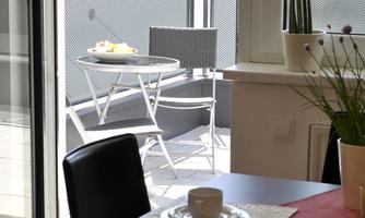 A modern dining room with a table and two coffee cups. Behind the glass sliding door, there is a small balcony with a table and chairs.
