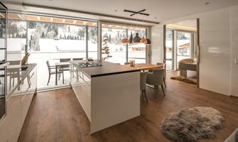 A modern kitchen with an open living area and large windows offering a view of the snow. The space is bright and inviting, featuring wooden floors and stylish furniture.