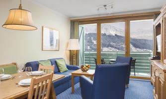 A cozy living room with blue furniture and a dining table. The large windows offer a view of the mountains.