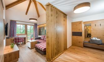 A cozy room with wooden decor, a sofa, and a dining table. Large windows offer a beautiful view of nature.