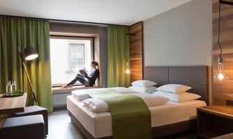 A modern hotel room with a large bed and a cozy window seat. A person sits relaxed and reads in the window nook.