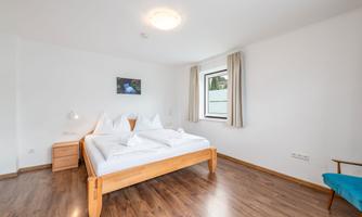 A bright bedroom with a large bed and natural wood flooring. The room has a large window and a cozy armchair.