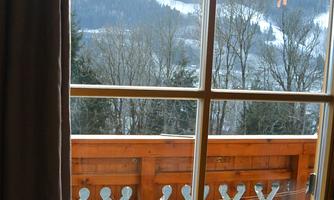 A view from a window onto a balcony with a wooden railing. In the background, snowy mountains and trees can be seen.