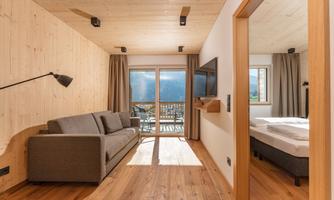 A modern room with wooden paneling, a cozy sofa, and large windows that provide plenty of light. In the background, a television and a bed are visible.