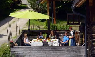 A group of five people is sitting relaxed on a terrace. They are enjoying a shared meal under a green sun umbrella.