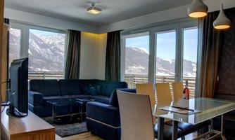 A modern living room with a blue couch and a dining area. Large windows offer a view of the snow-covered mountains.