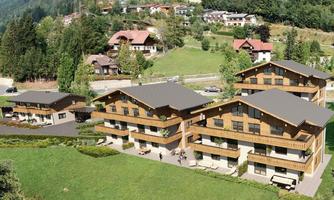 A modern residential complex with wooden buildings surrounded by green landscape. In the background, gentle hills and small houses are visible.