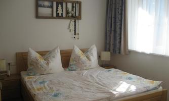 A bright bedroom with a large bed and pillows shaped like lace. Pictures and a plant decoration are hanging on the wall.