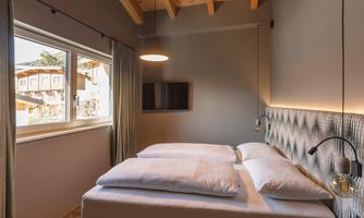 A modern bedroom with a large window that lets in daylight. The room is minimally furnished with a double bed and a flat-screen TV on the wall.