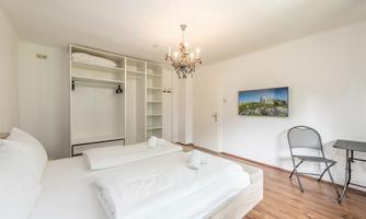 A modern furnished room with a double bed and wooden floor. A table, a chair, and a TV with a landscape picture complete the furnishings.
