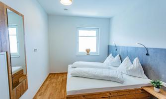 A modern bedroom with two single beds and a wooden frame. The walls are painted light, and the room has large windows as well as a wall mirror.