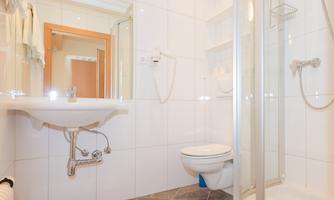 A modern bathroom with white tiles and a shower. In the foreground, a sink and a toilet area can be seen.