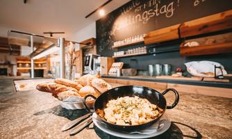 A rustic table with a pan of delicious food and a bowl of fresh bread. Wooden shelves and a cozy atmosphere can be seen in the background.