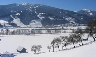 A winter landscape with snow-covered fields and hills. In the background, mountains and a clear blue sky are visible.