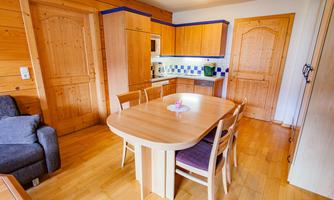 A cozy kitchen with wooden furniture and a large dining table. The walls are light-colored, and there is a seating area with a gray sofa.