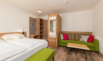 A modern room with a double bed and a cozy sofa. The decor is bright and friendly, with wood and green tones.