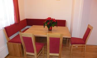 A cozy dining area with a wooden table and red chairs.  
There is a bench-like seating area against the wall and a blooming pot.