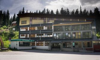 A modern building with large windows is situated in a mountainous area. Surrounded by forests, it presents an inviting sight.