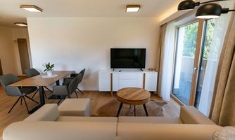 A modern living room with a comfortable sofa and a table. A television and large windows provide plenty of light.