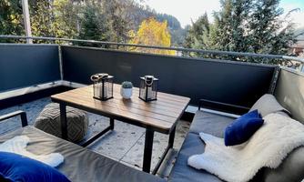 A cozy balcony with a wooden table and relaxing sofas. In the background, there are trees and a clear, fresh landscape.