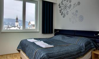 A modern bedroom with a large bed and a blue bedspread. Through the window, one looks out onto a snowy landscape and a church tower silhouette.