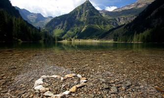 A clear lake with a heart made of stones in the foreground. In the background, majestic mountains and a blue sky can be seen.