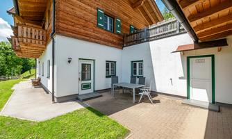 A cozy house with wooden cladding and green window frames. Outside, there is a dining table and a terrace surrounded by lawn.