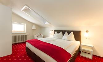 A cozy bedroom with a double bed and red accents. There is a window, a lamp, and a bedside table.
