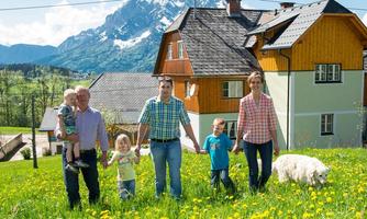 A family stands in a blooming field in front of a house with mountains in the background. They are holding hands and enjoying a sunny day.