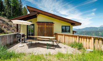 A cozy house in the mountains with a terrace and an outdoor table. In the background, green meadows and trees can be seen.