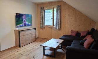A modern living room with a television, a sofa corner, and a wooden table. The walls are bright and the furniture creates a cozy atmosphere.
