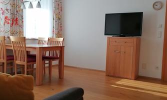 A bright living room with a dining table and wooden furniture. A television is on a cabinet against the wall.