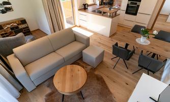 A modern living and dining room with a beige sofa, a round wooden table, and black chairs. The rooms are bright and friendly, featuring wooden floors and large windows.