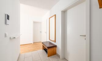 A bright hallway with white walls and a wooden floor. On the right side, there is a mirror and a wooden bench.