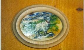 An oval picture of a goat in a green landscape. The background features mountains and a blue sky.
