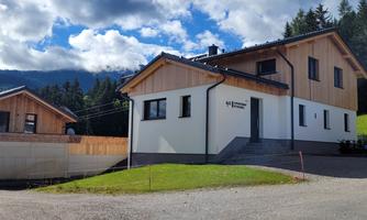 A modern house in a rural setting, surrounded by green meadows and mountains. The sky is partly cloudy.