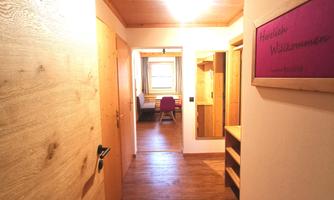 A bright corridor with wooden doors and an inviting design element. At the end of the corridor, there is a room with furniture visible.