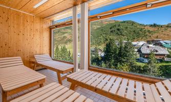 A modern relaxation area with wooden loungers and large windows. The view shows a green mountain landscape and a small village.