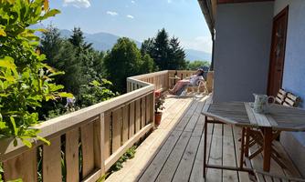 A sunny terrace with wooden railing and comfortable furniture. In the background, green trees and mountains are visible.