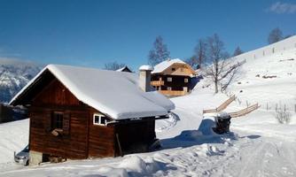 A snowy landscape with wooden chalets under a clear blue sky. The path leads through the snow to the cabins.