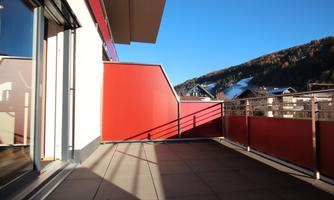 A modern balcony with a red railing and a view of the mountains. The sky is clear and blue.
