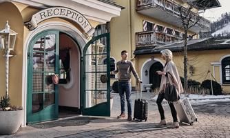 A reception area of a hotel with a woman and a man. The man greets the woman who is arriving with a suitcase.