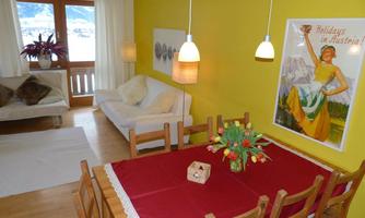A bright living room with yellow walls and a large dining table. The image shows a sofa, some plants, and a vintage poster.