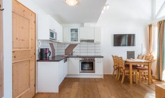 A modern kitchen with wooden flooring and light walls. A dining table with chairs is located on the side, while kitchen appliances are well arranged.