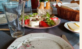 A set table with an empty plate, a glass of water, and a salad plate with tomatoes and fresh vegetables. In the background, there is a bottle and a basket of food.