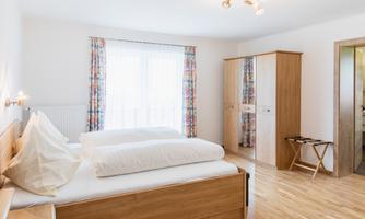 A bright bedroom with two beds and large windows. The furniture is made of wood and creates a cozy atmosphere.