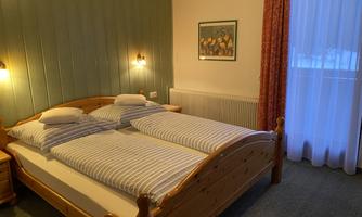 A cozy bedroom with a double bed and patterned cushions. The room has a warm atmosphere with soft lighting and a large window.
