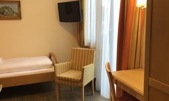 A simple hotel room with a single bed, an armchair, and a table. A television and large windows provide a bright living space.